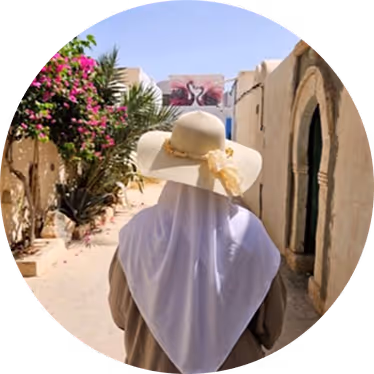 Une personne portant un foulard blanc et un chapeau de soleil beige marche dans une rue étroite et ensoleillée, bordée de fleurs et de portes cintrées.
